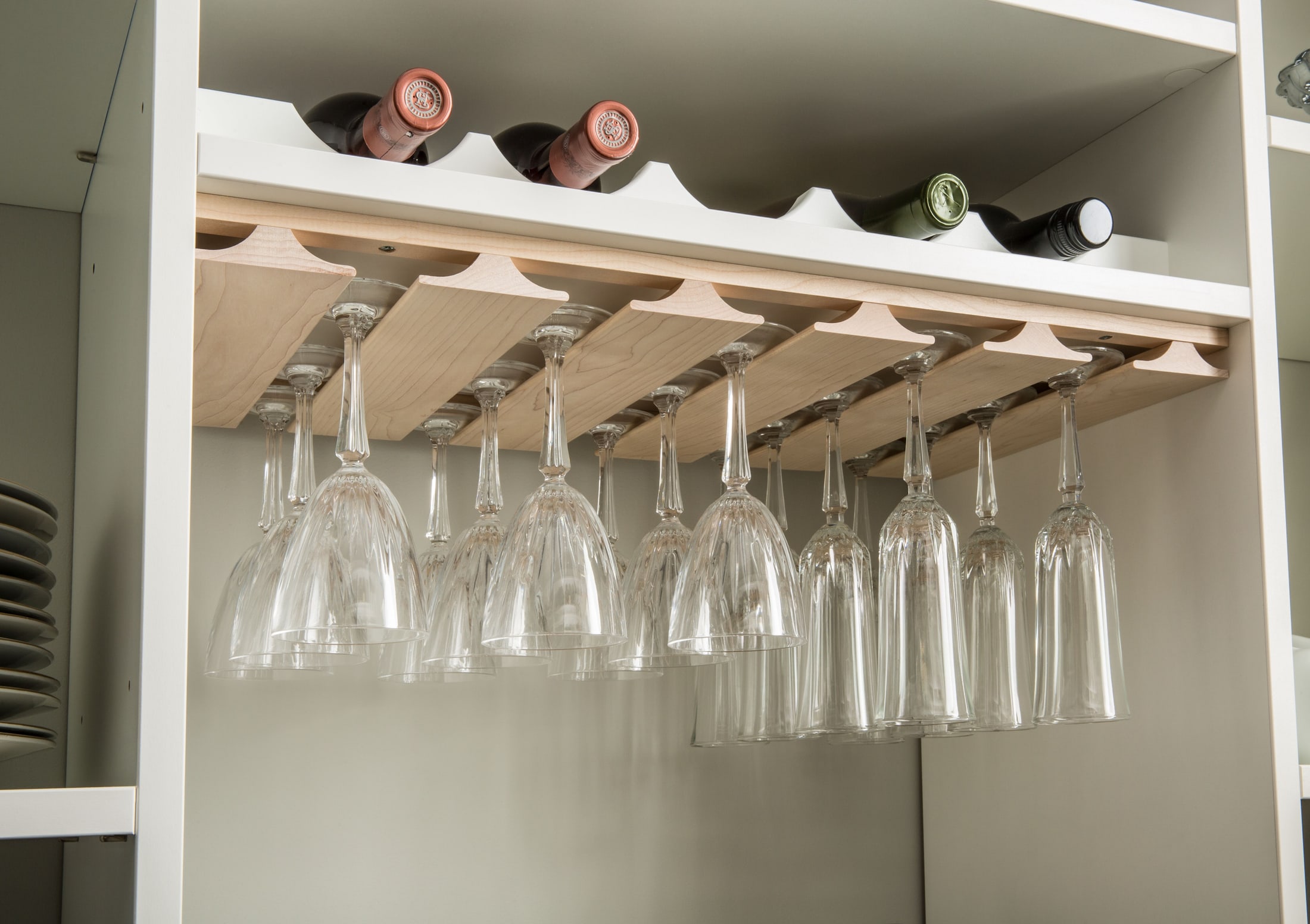 Maple under-shelf stemware rails holding rows of hanging wine glasses beneath a scalloped bottle rack in a custom pantry